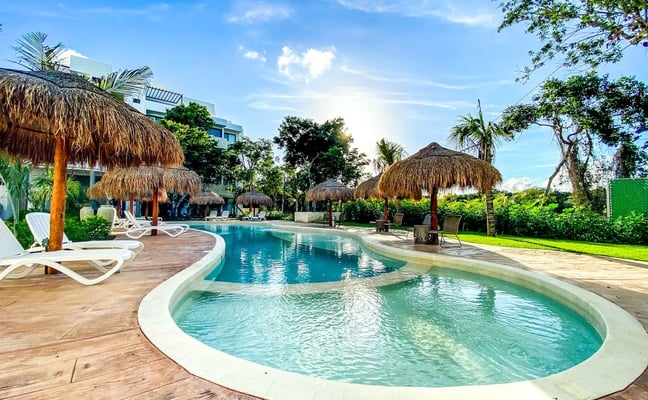 Área de piscina de resort con palapas de techo de palma, tumbonas, edificio moderno y vegetación tropical bajo un cielo azul